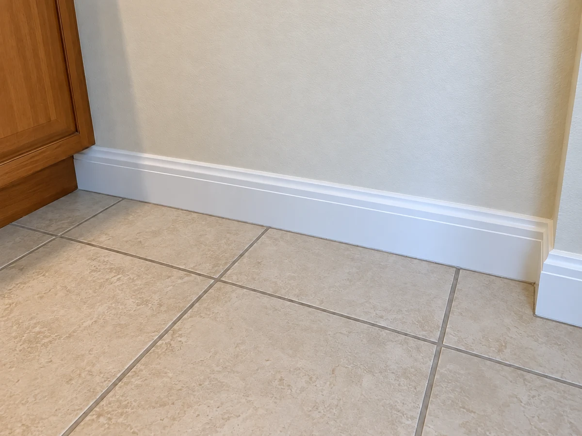After: the same baseboard and tiled floor corner with the dust and debris removed and clean grout lines, beside the wooden cabinet edge.