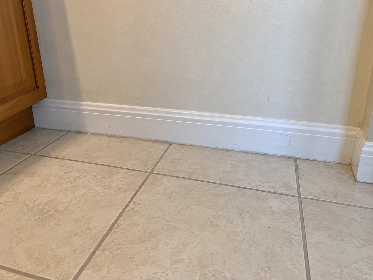 Before: a white baseboard and tiled floor corner with visible dust and debris along the baseboard and grout lines, beside a wooden cabinet edge.