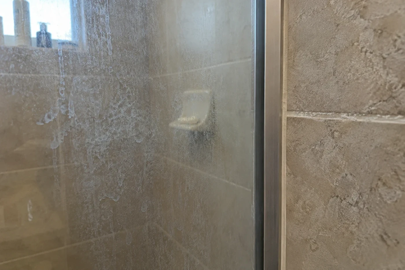 Soap scum and mineral build-up on a glass shower enclosure and along a tile grout line in a home bathroom.
