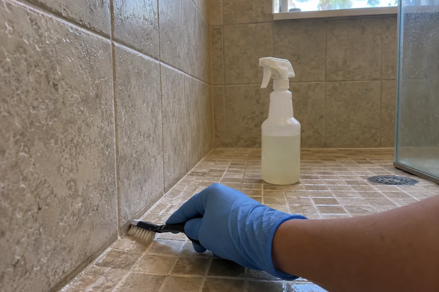A gloved hand using a small detail brush along a tile edge in a shower, with an unbranded spray bottle nearby.