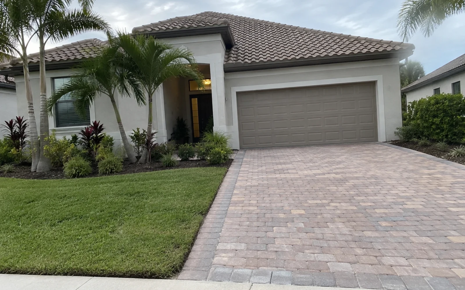 An ordinary consumer-phone snapshot of a newer single-storey Estero home with a paver driveway and young palms in flat daylight, with slightly imperfect framing.