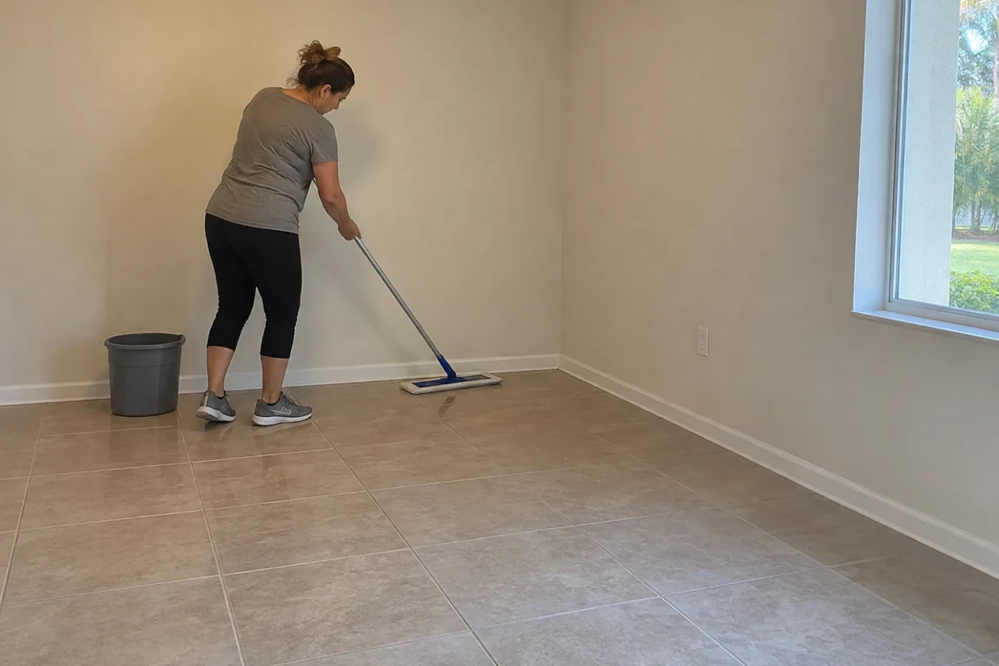 A person in plain clothes mopping along the floor edge of an empty room, with a bin and a mop beside them and no belongings in the room.